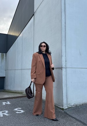 Mujer con traje marrón y blusa negra, sosteniendo un bolso oscuro, de pie sobre asfalto cerca de un gran edificio de concreto color gris claro.