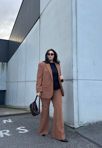 Mujer con traje marrón y blusa negra, sosteniendo un bolso oscuro, de pie sobre asfalto cerca de un gran edificio de concreto color gris claro.