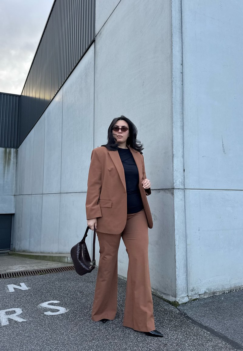 Woman wearing brown suit and black top, holding dark handbag, standing on asphalt near large light gray concrete building.