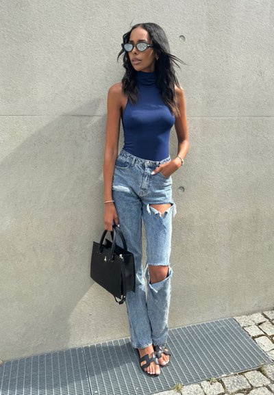 Top de cuello alto sin mangas color navy, jeans desgastados de color azul claro con rasgaduras en las rodillas, bolso negro y sandalias negras sobre una superficie gris texturizada.