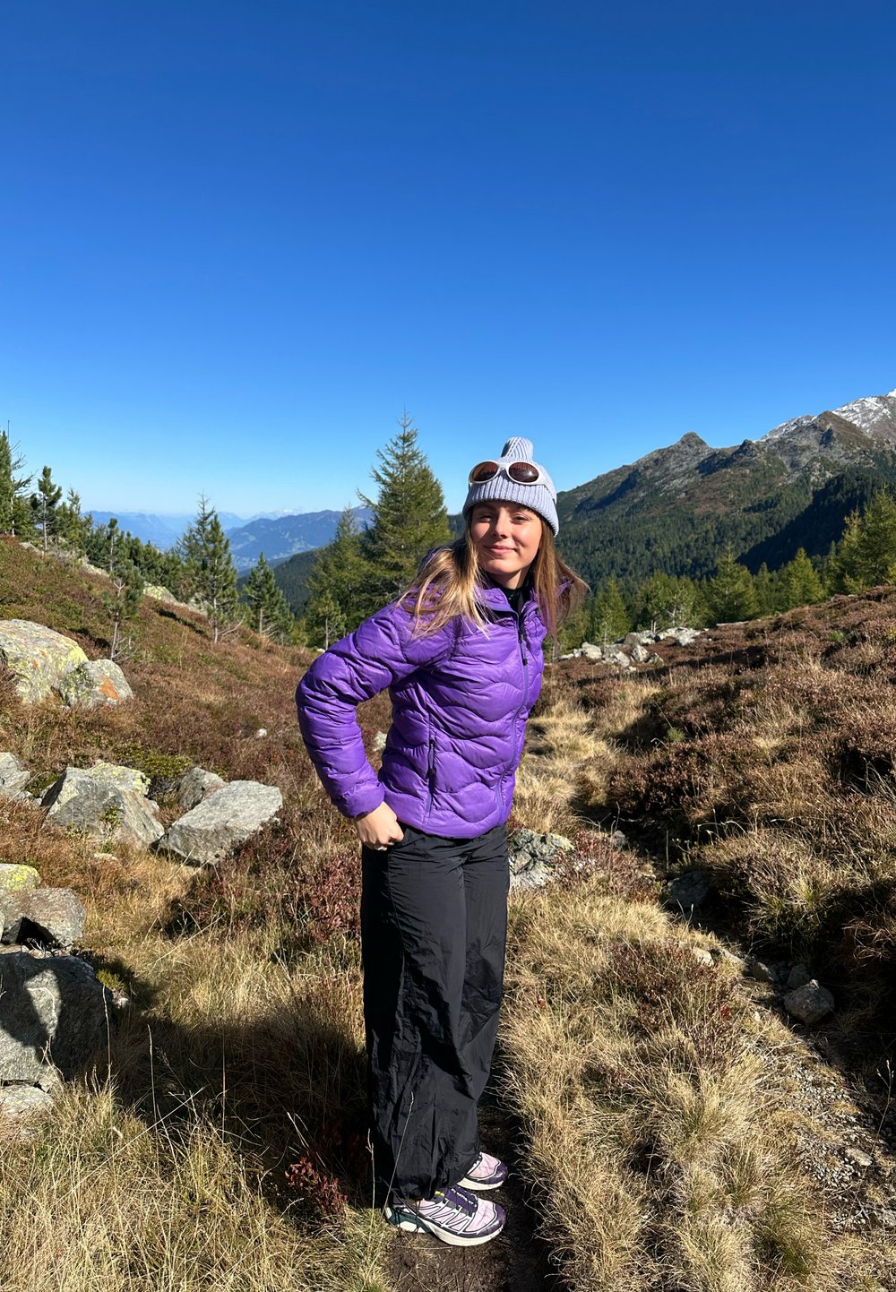 Giovane donna con giacca viola, pantaloni neri e berretto grigio che fa escursione su un sentiero di montagna circondato da alberi di pino e con cielo blu limpido.