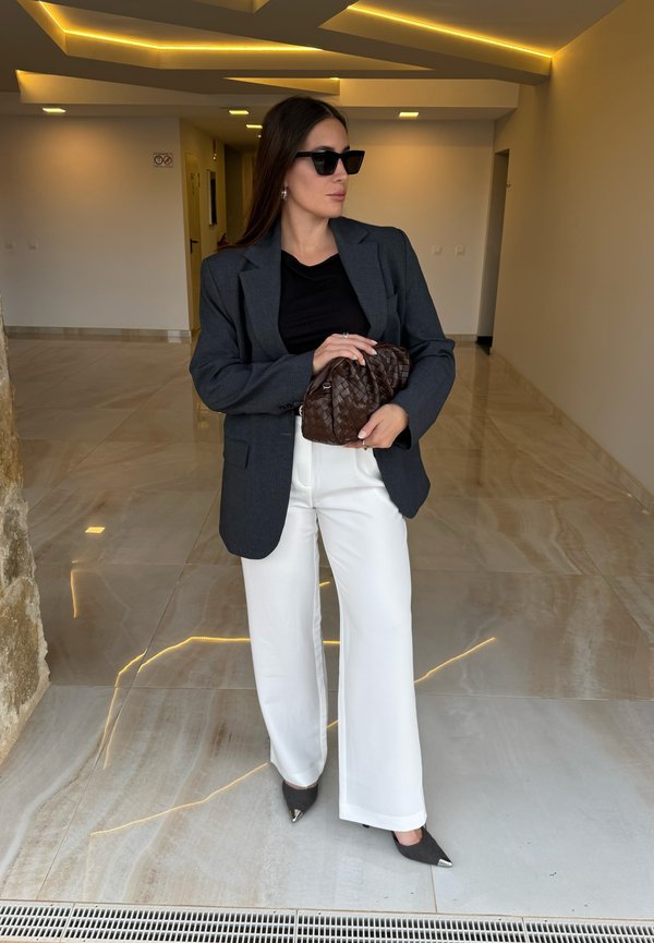 Grijze blazer, zwart shirt, witte wijde broekspijpen, donkere zonnebril, een bruine gevlochten clutch vasthoudend, staand op een glanzende marmeren vloer.
