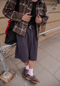 Jachetă maro cu carouri, cămașă neagră, pantaloni scurți gri, pantofi maroon cu șosete albe. Outfit-ul include un brâu negru și inele la degete.