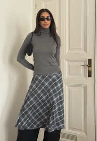 Femme portant des lunettes de soleil noires, un col roulé gris, une jupe à carreaux, tenant un petit sac noir, debout à l'intérieur près d'une porte blanche à panneaux.