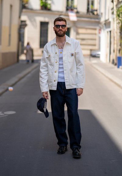 Chaqueta de algodón blanca con botones dorados, combinada con una camiseta sin mangas blanca y jeans azul oscuro. Gafas de sol oscuras y una gorra azul marino en la mano.