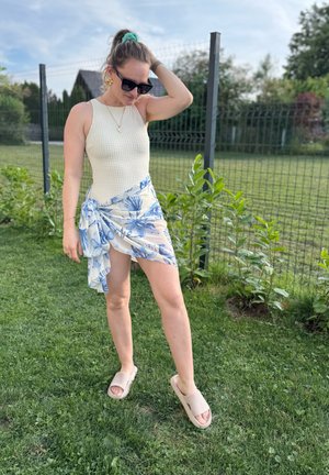 Bodysuit blanco y texturizado combinado con una falda envolvente floral de color azul claro. Lleva sandalias beige, de pie sobre un césped con una cerca de fondo.