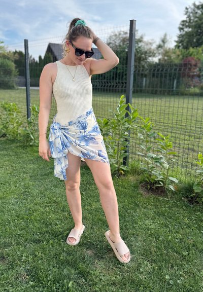 Bodysuit blanco y texturizado combinado con una falda envolvente floral de color azul claro. Lleva sandalias beige, de pie sobre un césped con una cerca de fondo.