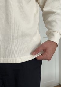 Pull en maille gaufrée de couleur crème, à manches longues, avec des poignets côtelés contrastants, légèrement ajusté à la taille, porté avec un pantalon sombre.