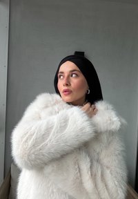 Abrigo de piel sintética blanca con textura esponjosa y ajuste suelto, con un amplio cuello y mangas suaves y voluminosas. Un pañuelo negro en la cabeza realza el estilo.