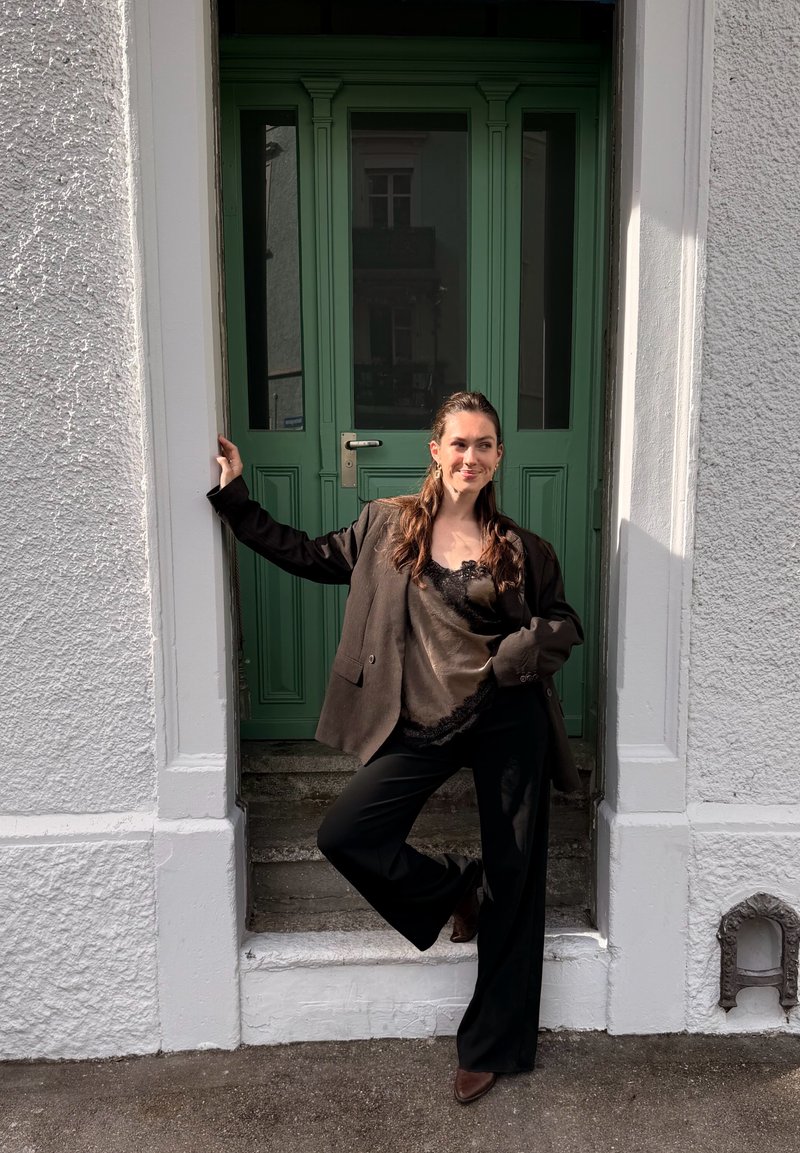 Femme en blazer foncé, top en satin et pantalon noir, s'appuie contre un cadre de porte vert avec une jambe levée sur des marches en béton.