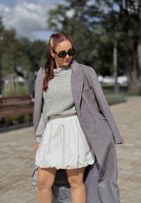 Palton lung gri peste un pulover tricotat gri și o fustă pufos albă. Ochelari de soare rotunzi negri. Texturi netede și culori moi. Cadru în aer liber.