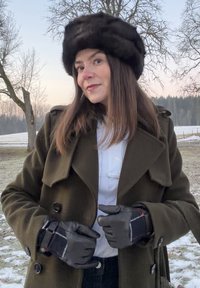 Γυναίκα με σκοτεινό γούνινο καπέλο, ελιάς πράσινο παλτό, λευκό πουλόβερ και μαύρα γάντια, στέκεται σε εξωτερικό χώρο σε χιονισμένο τοπίο με γυμνά δέντρα στο λυκόφως.