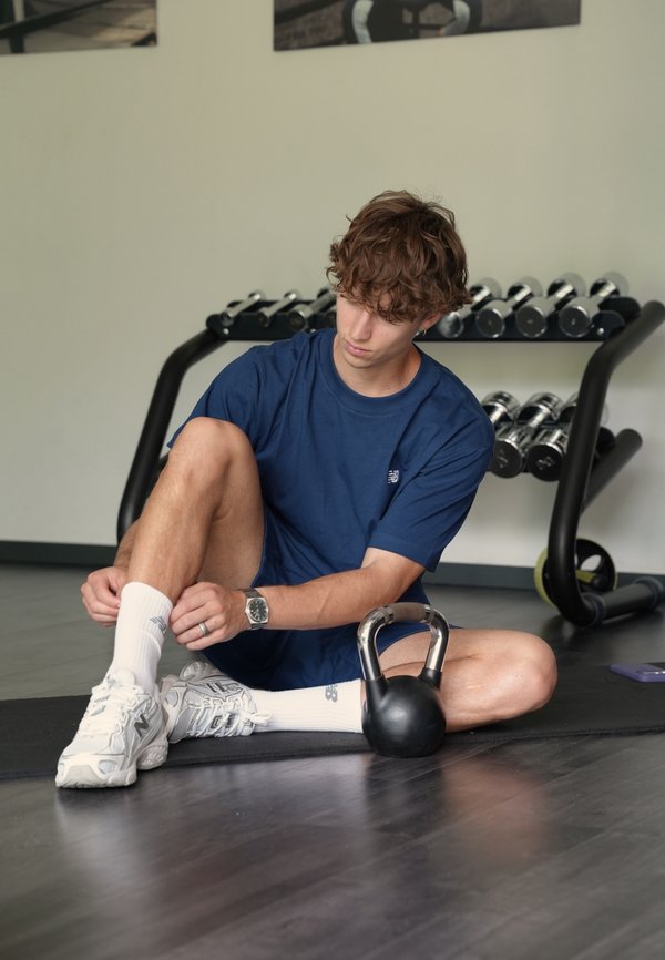 Mladý muž v modrom športovom oblečení sedí na telocvičnej podložke a upravuje si ponožku, v pozadí je kettlebell, činky a stojan.