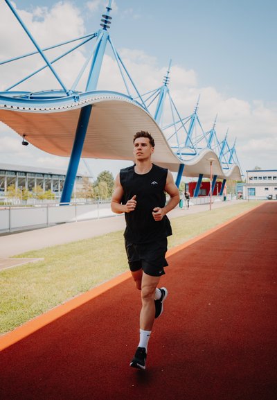 Top sin mangas negro y pantalones cortos, corriendo en una pista roja. Estructura arquitectónica moderna en azul y blanco en el fondo bajo un cielo despejado.