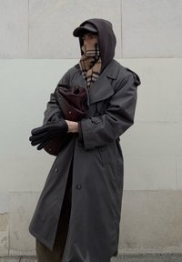 Long manteau trench gris, gants noirs, écharpe à carreaux et sac foncé ; personne portant une veste à capuche et une casquette se tient contre un mur clair.
