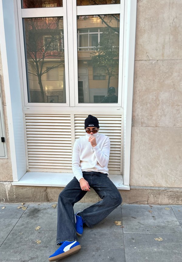 Vit ribbad stickad tröja, mörk vida jeans, blå sneakers med beige sula, svart mössa, runda solglasögon, sittande vid ett fönster.