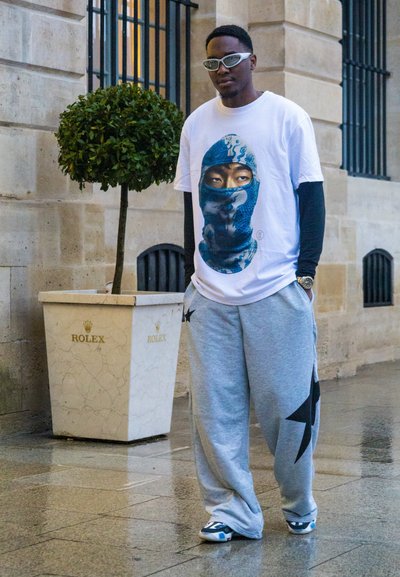 Camiseta blanca con un diseño gráfico azul que presenta una figura con capucha, combinada con pantalones de chándal grises con un patrón de estrellas negras y gafas de sol.