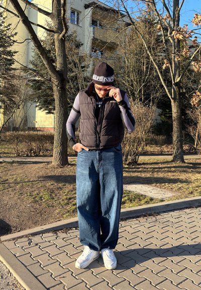 Chaleco de plumas negro, camisa de rayas de manga larga, vaqueros azules holgados y zapatillas blancas. Lleva un gorro negro con un logo. Ambiente exterior iluminado por el sol.