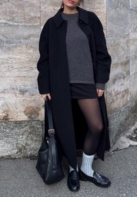 Γυναίκα με μακρύ μαύρο παλτό, γκρι πουλόβερ, μαύρη φούστα, μαύρο καλσόν, λευκές κάλτσες και μαύρα μοκασίνια, κρατώντας μαύρη τσάντα, στέκεται δίπλα σε πέτρινο τοίχο.