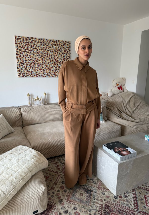 Mujer con pañuelo beige en la cabeza y ropa suelta marrón de pie en un salón con sofás beige, alfombra estampada, oso de peluche y arte en la pared.
