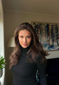 Młoda kobieta o długich brązowych włosach, ubrana w czarny golf, stojąca w pomieszczeniu przy oknie, za nią znajduje się lampa, roślina i abstrakcyjny obraz.