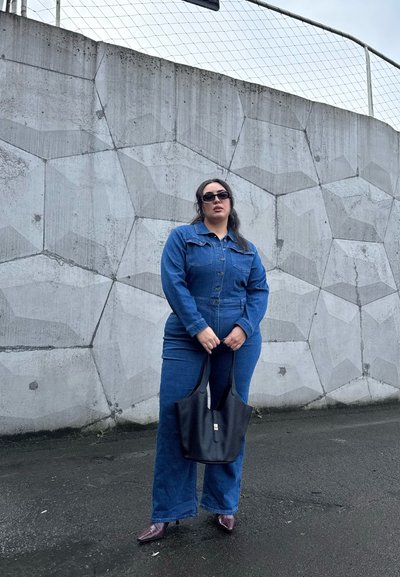 Mono de denim en azul oscuro con cierre de botones, combinado con un bolso tipo tote negro. Gafas de sol redondas y zapatos de tacón marrones completan el look.