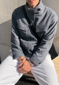 Hall hallitextuuriga jakk kõrge krae ja kahe eesmise taskuga, kantud valgete pükste kohal. Käevõru ja hõbedane käekell käes.