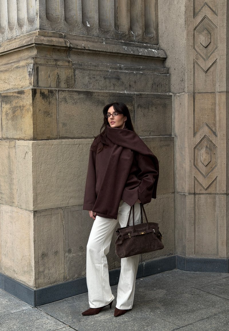 Oversized brown jumper with draped design, white wide-leg trousers, and brown suede handbag. Set against textured stone wall.
