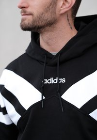 Črna majica s kapuco z belimi diagonalnimi črtami in vezenim logotipom. Izdelana iz mehkih materialov z žepom spredaj in nastavljivimi vrvicami.