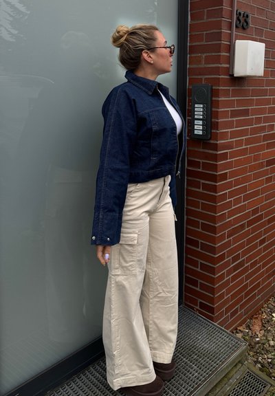 Chaqueta de mezclilla oscura corta, camisa blanca debajo, pantalones cargo anchos de color crema y botas de tobillo marrones. De pie junto a una pared de ladrillo y un intercomunicador.