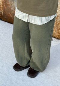 Pantalon large et ample de couleur vert olive avec une texture douce, assorti à une chemise rayée bleu clair et blanche ainsi qu'à des chaussures marron.