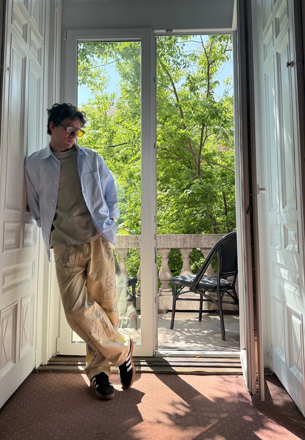 Világoskék ing, olíva zöld póló, bézs mintás nadrág, fekete sportcipő, egy nagy üvegajtó mellett áll, kívül zöld növényekkel.