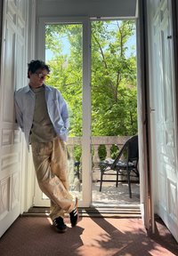 Világoskék ing, olíva zöld póló, bézs mintás nadrág, fekete sportcipő, egy nagy üvegajtó mellett áll, kívül zöld növényekkel.