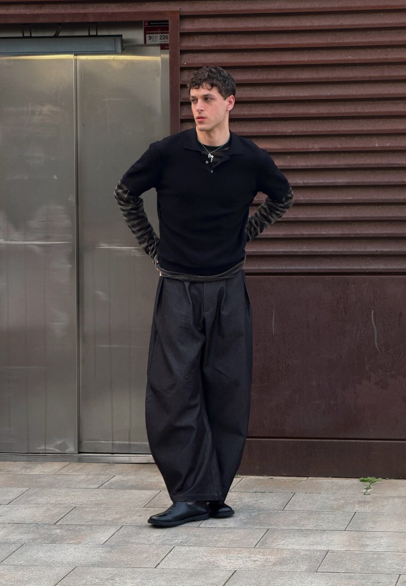 Jeune homme portant un pantalon noir à jambes larges, des hauts noirs superposés avec des manches à motifs, et des chaussures noires, debout sur le trottoir, les mains derrière le dos.