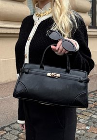 Ξανθιά γυναίκα με μαύρη και λευκή ζακέτα κρατά μαύρη δερμάτινη τσάντα και μαύρα γυαλιά ηλίου, στέκεται σε πλακόστρωτο δρόμο κοντά σε πέτρινο κτίριο.