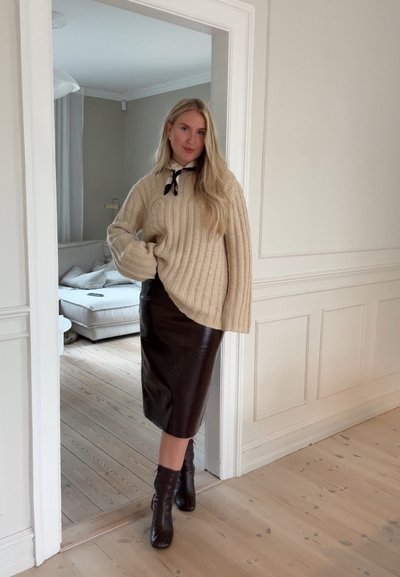 Suéter beige de canalé con mangas sueltas, falda midi de cuero negra y botas negras de tobillo, ambientado en una habitación de tonos neutros.