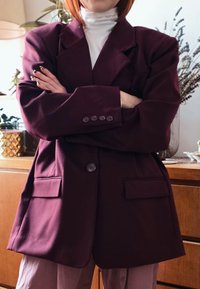 Persona con le braccia incrociate che indossa un blazer sovradimensionato di un viola scuro e un dolcevita bianco in una stanza con mobili in legno e piante.