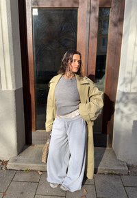 Trenci kaki, pulover gri și pantaloni sport gri deschis. Ținuta are un croi lejer și este completată cu o geantă cu model și adidași.
