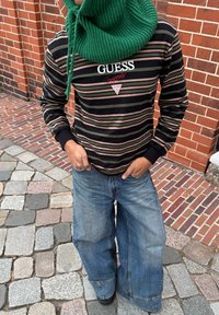 Osoba stoji na kaldrmisanoj ulici noseći prugastu Guess majicu, traperice širokih nogavica, crne cipele i veliki zeleni pleteni šal.