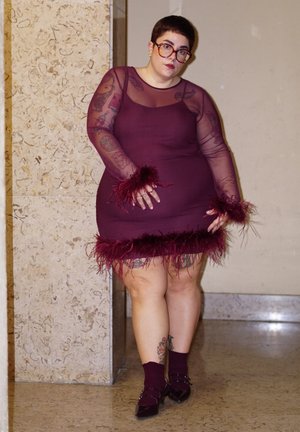 Persona con cabello corto y gafas, vestida con un vestido largo y transparente de manga larga color granate, con puños y dobladillo con plumas, de pie contra una pared de piedra.