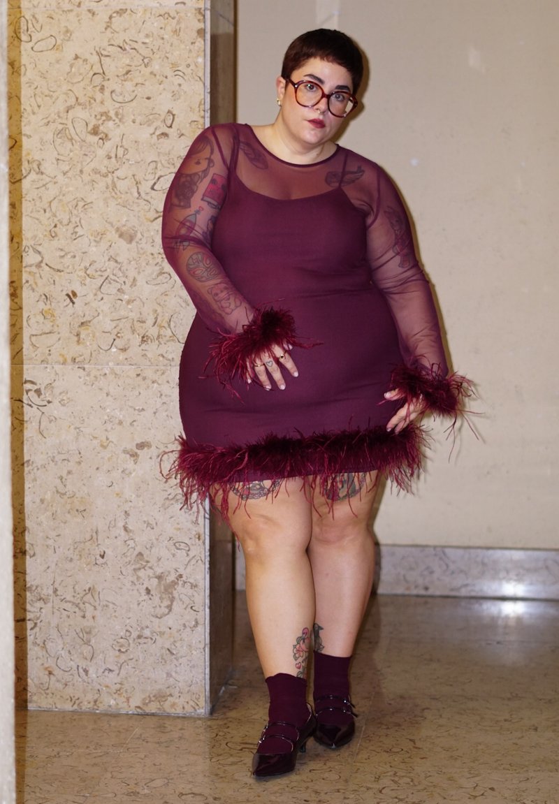 Person mit kurzen Haaren und Brille, gekleidet in ein durchsichtiges, langärmeliges, kastanienbraunes Kleid mit gefiederten Manschetten und Saum, stehend vor einer Steinmauer.