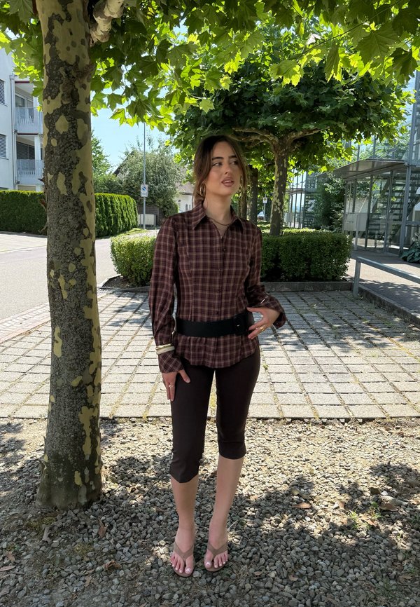 Kvinde iført en brun ternet skjorte, sorte korte bukser og sandaler, står under et løvtræ på en solrig fortov.