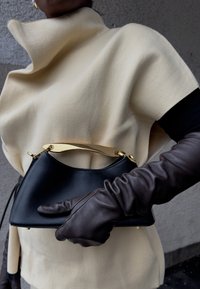 Personne portant une cape crème et de longs gants en cuir marron foncé tient un petit sac à main noir avec une poignée dorée sur un fond gris.