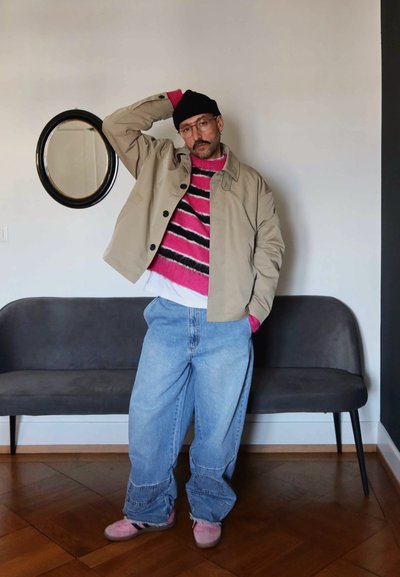 Chaqueta marrón, suéter a rayas rosa y negro, jeans azules holgados y zapatos rosas. De pie junto a un sofá gris y un espejo redondo en una pared blanca.