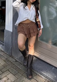 Falda mini marrón con volantes y un cinturón de cadena dorada, combinada con una camiseta blanca corta y botas de cuero negras hasta la rodilla.