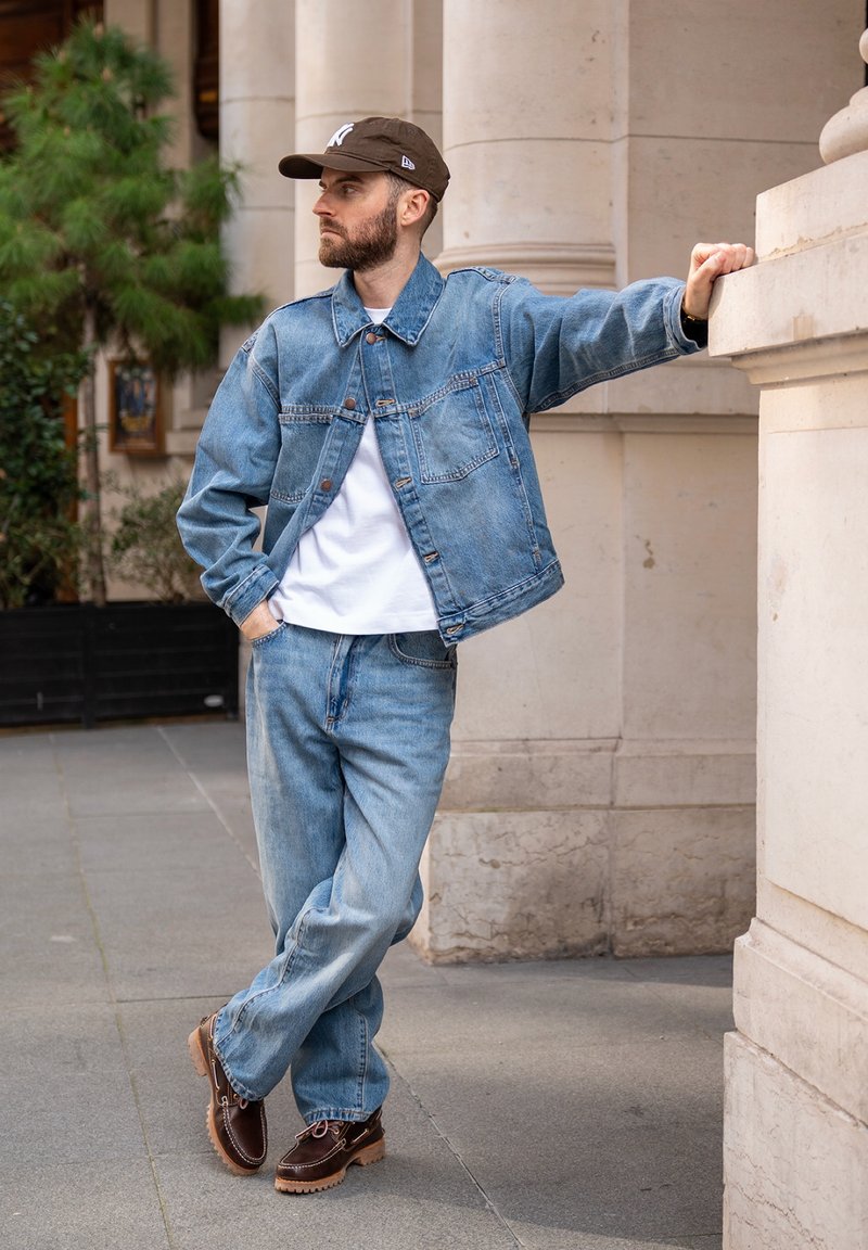 Homme en veste en jean et jean appuyé contre un pilier en pierre, portant un t-shirt blanc, une casquette marron et des chaussures marron sur un trottoir en ville.