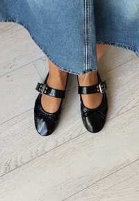 Czarne skórzane baleriny z zaokrąglonym noskiem, elastycznymi brzegami i podwójnymi zapinanymi paskami. Buty mają gładką teksturę i minimalistyczny design.