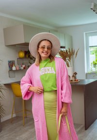 Pitkä vaaleanpunainen neulottu neule, vihreä paita, jossa on "KOMBUCHA" -painatus, limevihreä kynähame, leveähuuline beige hattu, keltaiset aurinkolasit, minimaalinen asustevalikoima.