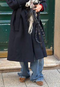 Personne en long manteau sombre et jean bleu ample tenant un petit chien noir et blanc duveteux avec une laisse.