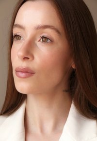 Nærbilde av et ansikt med brune øyne, glatt lys hud og langt brunt hår. Iført en beige blazer, med myke lepper og en nakketatovering.